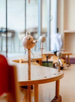 Handgefertigtes Montessori-Holzspielzeug steht auf einem halbrunden Tisch in einem hellen, freundlichen Klassenraum. Die detailreichen Holzfiguren laden Kinder zum kreativen Spielen ein. Im Hintergrund sorgen gro&szlig;e Fenster und dezentes Mobiliar f&uuml;r eine angenehme Lernatmosph&auml;re.