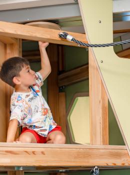 Ein kleines Kind erkundet neugierig einen h&ouml;lzernen Kletterbereich in einem Indoor-Spielplatz. Das Kind mit bunter Kleidung h&auml;lt sich an einer Stange fest und blickt auf die n&auml;chste Herausforderung. Die Szene vermittelt Freude, Aktivit&auml;t und Entdeckerlust in sicherer Umgebung.