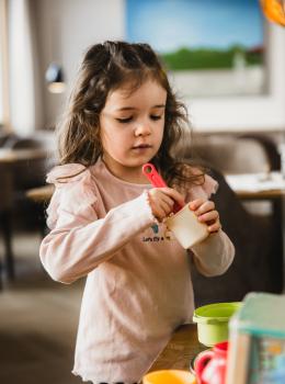 Ein junges M&auml;dchen steht an einem Tisch und spielt konzentriert mit buntem Spielgeschirr. Sie h&auml;lt einen kleinen Messl&ouml;ffel in der Hand und taucht ihn in einen Becher. Im Hintergrund ist ein gem&uuml;tlicher, heller Raum mit modernen M&ouml;beln sichtbar. Die Szene vermittelt eine freundliche und kreative Atmosph&auml;re.