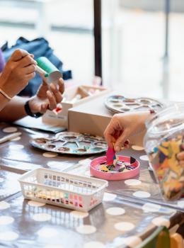 Mehrere H&auml;nde gestalten gemeinsam bunte Bastelarbeiten mit verschiedenen Materialien. Die Bastelutensilien liegen auf einem gepunkteten Tisch, w&auml;hrend im Hintergrund helle Fenster Licht spenden. Die Szene zeigt eine einladende, kreative Atmosph&auml;re, ideal f&uuml;r Kinder und Familienprojekte.