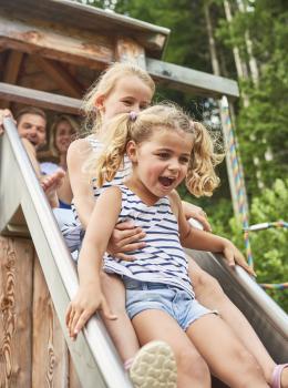 Fr&ouml;hliche Kinder gleiten gemeinsam eine Holzrutsche hinunter, umgeben von gr&uuml;nen B&auml;umen in einer naturnahen Umgebung. Die helle Sommerstimmung und das Lachen der Kinder vermitteln pure Lebensfreude und ein authentisches Naturerlebnis im Freien.