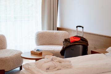 Ein helles, einladendes Hotelzimmer mit zwei weichen Sesseln, einem kleinen Holztisch und einem großen Bett, auf dem ein Bademantel liegt. Ein Koffer mit Jacke und Mütze steht bereit, um das komfortable Ambiente zu unterstreichen. Ideal für einen entspannten Aufenthalt.