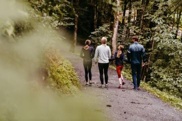 Eine Familie spaziert gemeinsam auf einem naturbelassenen Waldweg, umgeben von dichtem Grün und hohen Bäumen. Die entspannte Atmosphäre lädt zu gemeinsamen Erlebnissen in der Natur ein. Sanftes Licht unterstreicht die friedliche Stimmung dieser Wanderung im Freien.