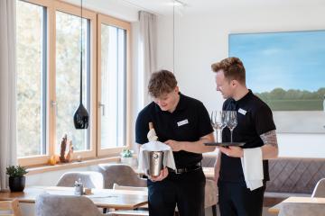 Zwei freundliche Servicekr&auml;fte im modernen Restaurant bereiten den Weinservice vor. Einer h&auml;lt eine gek&uuml;hlte Weinflasche mit Serviette, der andere serviert Gl&auml;ser auf einem Tablett. Helles Tageslicht f&auml;llt durch gro&szlig;e Fenster und sorgt f&uuml;r eine einladende Atmosph&auml;re am gedeckten Tisch.