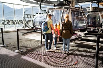 Eine Familie mit zwei Kindern bereitet sich auf den Einstieg in eine moderne Seilbahnkabine vor. Sonnenlicht durchflutet die helle Station, im Hintergrund sind Berge sichtbar. Die Szene vermittelt Vorfreude auf ein gemeinsames Abenteuer in der Natur.