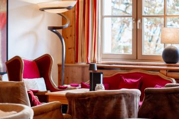 Eine stilvolle Lounge mit bequemen Sesseln und Sofas in warmen Rottönen lädt zum Verweilen ein. Großzügige Fenster lassen Tageslicht herein und bieten einen beruhigenden Ausblick. Die dekorativen Kissen, moderne Stehlampe und Akzentbeleuchtung schaffen eine behagliche, einladende Atmosphäre.