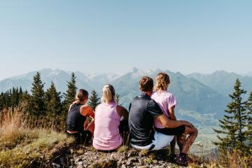 Vier Personen genie&szlig;en gemeinsam eine Pause auf einem Berggipfel und blicken auf die beeindruckenden Alpen. Umgeben von gr&uuml;nen B&auml;umen und sonniger Natur vermittelt das Bild Entspannung, Abenteuerlust und famili&auml;ren Zusammenhalt in malerischer Berglandschaft.