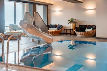 Ein stilvoller Innenpool mit einer glänzenden Edelstahlrutsche sorgt für Spaß und Entspannung. Im Hintergrund laden gemütliche Loungesessel und sanfte Beleuchtung zum Verweilen ein. Die klare Wasseroberfläche spiegelt die freundliche Atmosphäre des Wellnessbereichs wider.