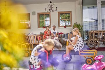Auf einer sonnigen Terrasse entspannen zwei Erwachsene auf einer Holzbank, w&auml;hrend Kinder auf bunten Spielzeugen fr&ouml;hlich spielen. Die Szene vermittelt ein lebendiges, famili&auml;res Ambiente mit liebevollen Details und einer gem&uuml;tlichen, einladenden Atmosph&auml;re. Ideal f&uuml;r Familienurlaub und gemeinsame Momente.