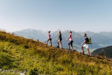 Eine vierköpfige Familie wandert bei strahlendem Sonnenschein entlang eines alpinen Wiesenhangs. Im Hintergrund türmen sich majestätische Berggipfel unter klarem Himmel. Die Szenerie strahlt Aktivität, Naturerlebnis und Sommerfreude aus – ideal für Outdoor-Fans.