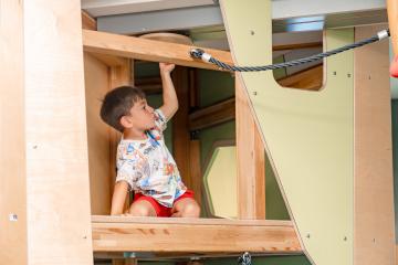 Ein kleines Kind erkundet neugierig einen hölzernen Kletterbereich in einem Indoor-Spielplatz. Das Kind mit bunter Kleidung hält sich an einer Stange fest und blickt auf die nächste Herausforderung. Die Szene vermittelt Freude, Aktivität und Entdeckerlust in sicherer Umgebung.