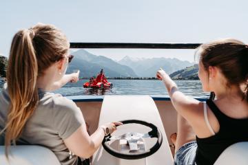 Zwei junge Frauen genießen eine Fahrt auf dem Tretboot bei strahlendem Sonnenschein. Sie zeigen auf ein weiteres Tretboot in der Ferne, umgeben von klaren Wasser und imposanten Berglandschaften. Die entspannte Atmosphäre lädt zum Entdecken und Verweilen ein.
