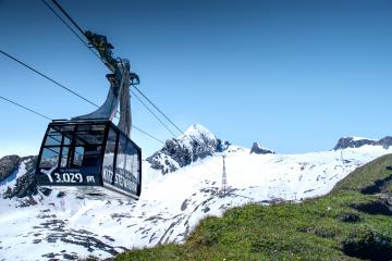 Eine moderne Seilbahngondel schwebt über die schneebedeckten Hänge des Kitzsteinhorns in den Alpen. Der blaue Himmel und das markante Bergpanorama schaffen eine beeindruckende Kulisse. Die Gondel bietet einen Panoramablick auf das alpine Winterwunderland und grüne Almwiesen im Vordergrund.