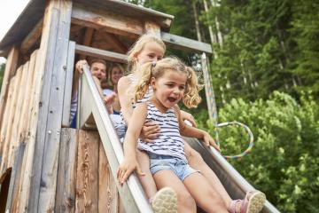 Fröhliche Kinder gleiten gemeinsam eine Holzrutsche hinunter, umgeben von grünen Bäumen in einer naturnahen Umgebung. Die helle Sommerstimmung und das Lachen der Kinder vermitteln pure Lebensfreude und ein authentisches Naturerlebnis im Freien.