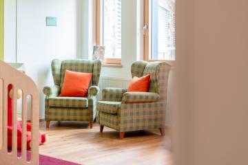 Zwei bequeme Polstersessel mit kariertem Muster und orangefarbenen Kissen stehen nebeneinander in einem hellen Raum mit Holzfußboden. Tageslicht fällt durch große Fenster und sorgt für eine freundliche, einladende Atmosphäre. Perfekt zum Entspannen und Wohlfühlen.