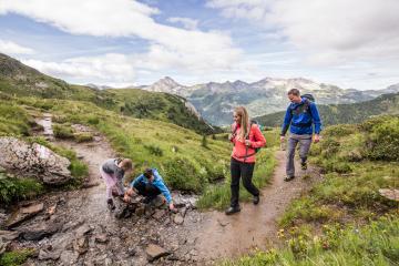 Eine Familie genießt eine Wanderung auf einem Gebirgspfad mit weitem Blick auf grüne Berge und eindrucksvolle Gipfel. Kinder spielen an einem kleinen Bach am Wegesrand, während Erwachsene aufmerksam begleiten. Die Szenerie vermittelt Naturverbundenheit, Abenteuerlust und entspannte Familienzeit im Freien.