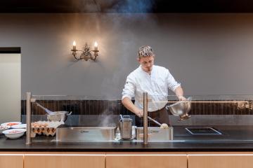 Ein Koch steht in einer modernen, offenen Küche und bereitet konzentriert Eierspeisen zu. Vor ihm liegen frische Eier und Küchenutensilien bereit. Der aufsteigende Dampf und die warme Beleuchtung schaffen eine einladende, professionelle Atmosphäre.
