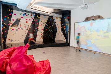 Ein heller Indoor-Spielraum mit einer farbenfrohen Kletterwand und gro&szlig;er interaktiver Projektionsfl&auml;che. Zwei Kinder sind im Raum aktiv, w&auml;hrend auff&auml;llige pinkfarbene Sitzs&auml;cke eine gem&uuml;tliche Ecke bilden. Die Kombination aus Bewegung und multimedialer Unterhaltung bietet vielseitigen Spielspa&szlig;.