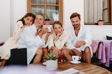 Eine gl&uuml;ckliche Familie genie&szlig;t gemeinsam Zeit auf einer sonnigen Terrasse. Alle tragen gem&uuml;tliche Badem&auml;ntel, trinken hei&szlig;e Getr&auml;nke und schauen entspannt in die Kamera. Die gem&uuml;tliche Atmosph&auml;re wird durch Pflanzen und sanfte Farben im Hintergrund unterstrichen.