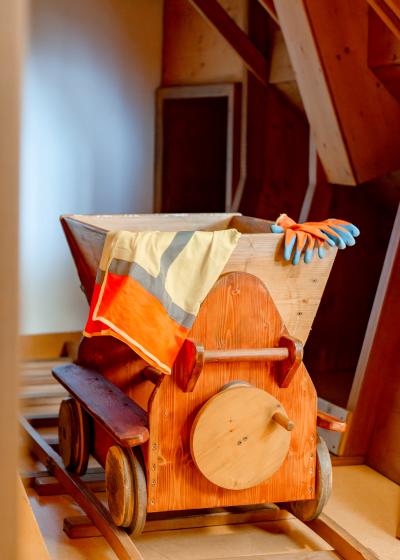 Ein liebevoll gefertigter Holz-Minenwagen steht auf Gleisen in einem kreativ gestalteten Kinderspielbereich. Ein farbenfroher Umhang und Handschuhe liegen &uuml;ber dem Wagen, was zum fantasievollen Spielen einl&auml;dt. Die warme Holzarchitektur sorgt f&uuml;r eine gem&uuml;tliche Atmosph&auml;re.