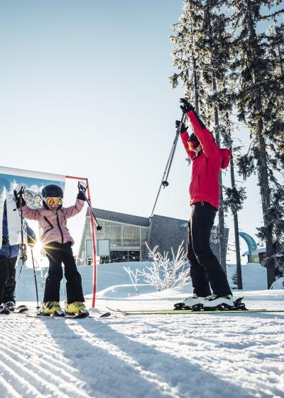 Ein Kind lernt mit Unterst&uuml;tzung eines Erwachsenen das Skifahren auf einer sonnigen, frisch pr&auml;parierten Piste. Die Szene zeigt Spa&szlig; und gemeinsame Aktivit&auml;t im familienfreundlichen Skigebiet, eingerahmt von verschneiten B&auml;umen und klarer Winterluft.