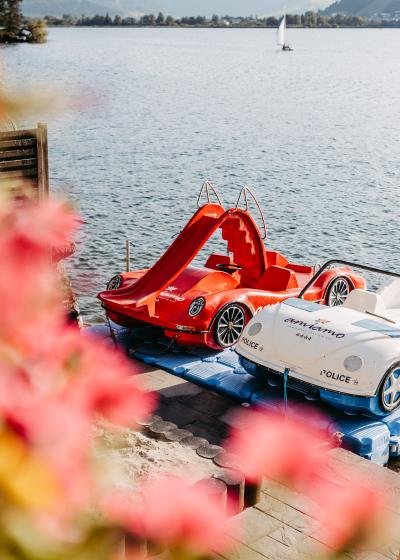 Zwei auffällige Tretboote im Sportwagendesign, eines rot mit Rutsche und eines weiß, liegen an einem Steg am ruhigen Seeufer. Im Vordergrund bunte Blumen, am Steg eine Person in Badebekleidung. Die Szene vermittelt Sommerstimmung und Freizeitspaß am Wasser.