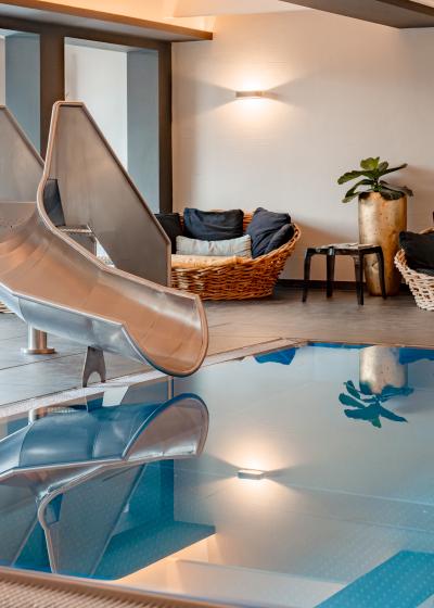 Ein stilvoller Innenpool mit einer gl&auml;nzenden Edelstahlrutsche sorgt f&uuml;r Spa&szlig; und Entspannung. Im Hintergrund laden gem&uuml;tliche Loungesessel und sanfte Beleuchtung zum Verweilen ein. Die klare Wasseroberfl&auml;che spiegelt die freundliche Atmosph&auml;re des Wellnessbereichs wider.