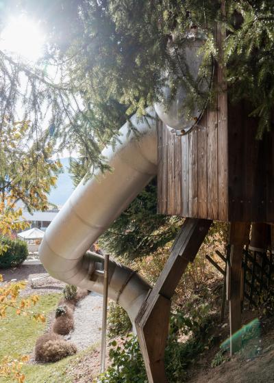 Eine gro&szlig;e, silberne Rutsche f&uuml;hrt aus einem h&ouml;lzernen Baumhaus und schl&auml;ngelt sich durch gr&uuml;ne Baumwipfel nach unten. Sonnenlicht gl&auml;nzt durch das Bl&auml;tterdach und verleiht der Szenerie eine verspielte und einladende Atmosph&auml;re. Perfekt f&uuml;r ein Abenteuer im Freien.