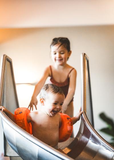 Zwei fröhliche Kinder genießen gemeinsam eine Rutschpartie im Innenbereich eines Schwimmbads. Das vordere Kind trägt orange Schwimmflügel und lacht, während das ältere Kind dicht dahinter unterstützend begleitet. Die Szenerie wirkt lebendig und vermittelt pure Badefreude.