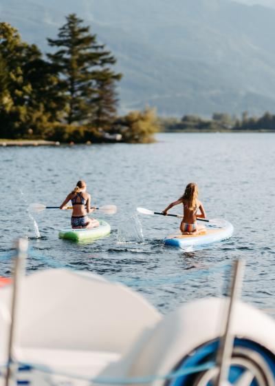 Zwei Personen genießen eine entspannte Stand-Up-Paddling-Tour auf einem klaren See, umgeben von einer malerischen Bergkulisse und grünen Bäumen. Im Vordergrund sind Tretboote zu sehen, während das ruhige Wasser und die idyllische Natur eine friedliche Atmosphäre schaffen.