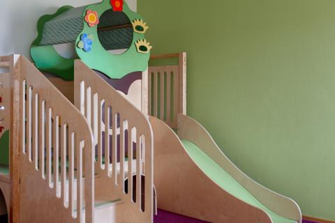 Ein moderner Indoor-Spielbereich mit einer kindgerechten Holzrutsche und einem bunten Kletterelement in Form eines Baumes. Die sanften Grün- und Holztöne schaffen eine freundliche Atmosphäre. Ideal zum sicheren Spielen und Klettern für Kleinkinder in Innenräumen.
