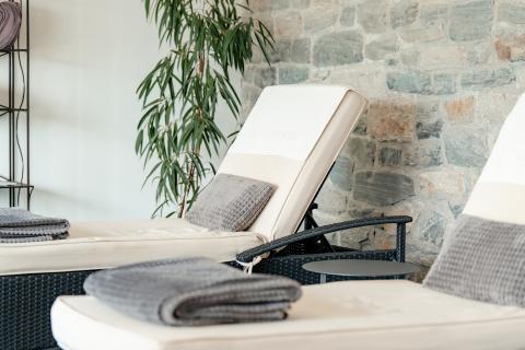 Zwei komfortable Relaxliegen mit weichen, grauen Handtüchern und Kissen stehen vor einer stilvoll gemauerten Steinwand. Eine grüne Pflanze sorgt für ein harmonisches Ambiente und lädt zum Wohlfühlen in entspannter Atmosphäre ein.