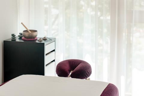 Ein heller, freundlich eingerichteter Massageraum mit einer bequemen Massageliege und weichem Tageslicht, das durch weiße Vorhänge fällt. Im Hintergrund steht eine Kommode mit einer Klangschale und harmonisch arrangierten Steinen – ideal für Erholung und Wohlbefinden.