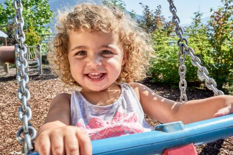 Ein fr&ouml;hliches Kleinkind mit lockigem Haar schaukelt auf einem Spielplatz an einem sonnigen Tag. Das Kind lacht in die Kamera, umgeben von gr&uuml;nen B&uuml;schen und B&auml;umen. Das Bild vermittelt Freude und unbeschwerte Kindheit im Freien.