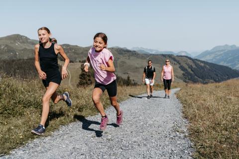 Zwei Kinder rennen fr&ouml;hlich einen Schotterweg entlang, w&auml;hrend im Hintergrund zwei Erwachsene gem&uuml;tlich spazieren. Die Szene spielt inmitten einer idyllischen Alpenlandschaft, umgeben von gr&uuml;nen H&uuml;geln und blauem Himmel &ndash; ideale Bedingungen f&uuml;r gemeinsame Familienaktivit&auml;ten in der Natur.
