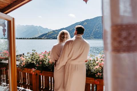 Ein Paar im Bademantel genießt entspannt die Aussicht vom Balkon auf einen idyllischen See, umgeben von farbenfrohen Blumen. Im Hintergrund erheben sich majestätische Berge unter klarem Himmel. Die warme Atmosphäre lädt zum Wohlfühlen und Verweilen ein.