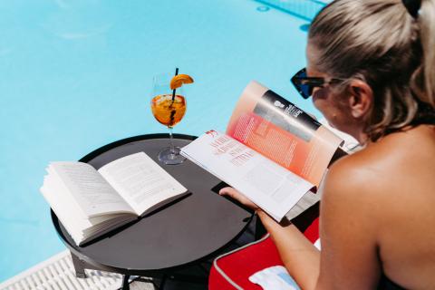 Eine Frau genießt einen sonnigen Tag am Pool, liest entspannt ein Magazin und hat ein weiteres Buch sowie einen erfrischenden Cocktail mit Orangenscheibe auf dem kleinen Tisch neben sich. Die Szene vermittelt Urlaubsfeeling und entspannte Sommeratmosphäre.