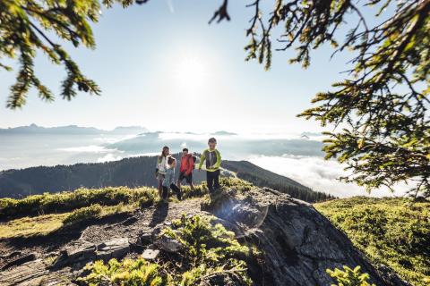 Eine fr&ouml;hliche Familie genie&szlig;t eine Wanderung auf einem sonnigen Bergr&uuml;cken, umgeben von gr&uuml;nen B&auml;umen und strahlendem Himmel. Sanfte Nebelfelder bedecken das Tal und schaffen eine idyllische Aussicht, w&auml;hrend die Sonnenstrahlen f&uuml;r eine lebendige, einladende Stimmung sorgen.