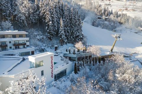 Das amiamo Hotel liegt eingebettet in eine verschneite Wald- und Berglandschaft direkt an einer Skipiste. Das moderne Gebäude mit markanter Beschriftung bietet einen perfekten Ausgangspunkt für Wintersport und Erholung im Schnee. Die schneebedeckten Bäume und die klare Winterluft schaffen eine gemütliche, idyllische Atmosphäre.