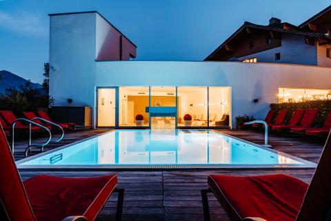 Moderner Außenpool auf einer stilvollen Holzterrasse, umgeben von roten Liegestühlen. Die warme Beleuchtung des Gebäudes spiegelt sich im klaren Wasser und sorgt für ein einladendes Ambiente am Abend. Perfekt für entspannte Sommernächte im Freien.