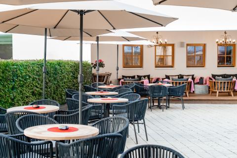 Modern eingerichtete Terrasse mit stilvollen Sitzplätzen, schattenspendenden Sonnenschirmen und liebevoll gedeckten Tischen. Im Hintergrund laden gemütliche Lounge-Bereiche und große Fenster zu entspannten Stunden ein. Perfekt für geselliges Beisammensein in einem freundlichen, einladenden Ambiente.