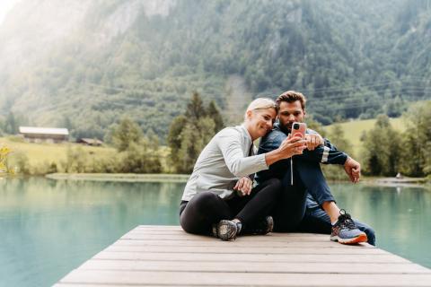 Ein gl&uuml;ckliches Paar sitzt entspannt auf einem Holzsteg am klaren See und macht gemeinsam ein Selfie. Sanfte Natur im Hintergrund mit gr&uuml;nen H&uuml;geln, W&auml;ldern und Bergh&auml;ngen schafft eine ruhige, idyllische Atmosph&auml;re. Perfekter Moment f&uuml;r unvergessliche Erinnerungen in alpiner Landschaft.