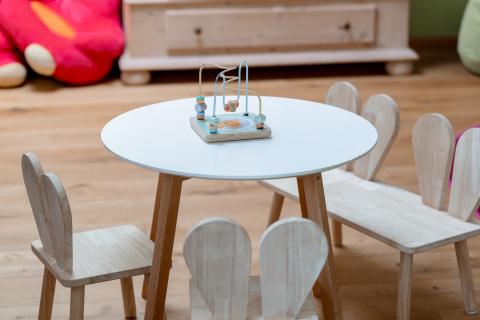Ein runder, weißer Kindertisch mit vier hellen Holzstühlen in Herzform steht auf einem Holzboden. In der Mitte des Tisches befindet sich ein Motorikspielzeug. Die freundliche Umgebung eignet sich ideal für Kinderaktivitäten und fördert spielerisches Lernen.
