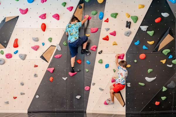 Zwei Kinder klettern an einer bunten Boulderwand mit verschiedenen farbigen Griffen und Tritten. Die Wand bietet abwechslungsreiche Routen und eine spannende Herausforderung in einer sicheren Indoor-Umgebung. Perfekt zum Spielen, Ausprobieren und Fördern motorischer Fähigkeiten.