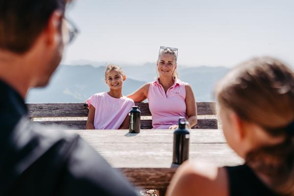 Eine lachende Mutter mit ihrem Kind genie&szlig;t den Tag auf einer Bergh&uuml;tte, w&auml;hrend Vater und Tochter ihnen gegen&uuml;ber sitzen. Im Hintergrund erstreckt sich eine beeindruckende Alpenlandschaft. Wasserflaschen auf dem Holztisch runden die gem&uuml;tliche Stimmung ab.