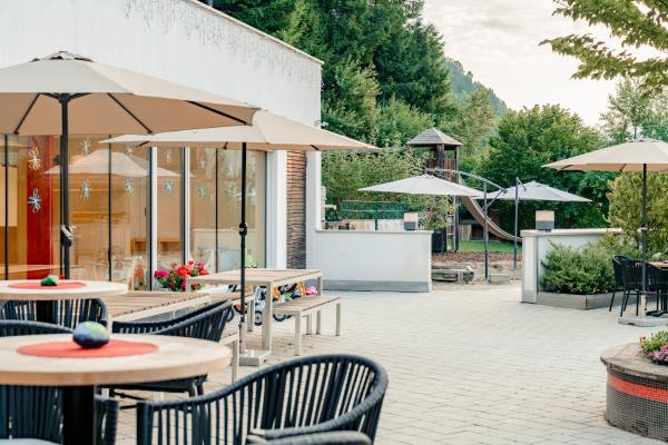 Die helle Sonnenterrasse bietet moderne Sitzgelegenheiten unter großen Sonnenschirmen und lädt zum entspannten Verweilen ein. Im Hintergrund sind ein Spielplatz und üppiges Grün zu sehen – ideal für Familien und Genießer. Die einladende Atmosphäre sorgt für Erholung im Freien.