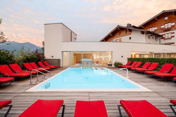 Exklusiver Außenpool mit Panoramablick Moderner Außenpool mit klaren Linien, umgeben von stilvollen roten Liegestühlen auf Holzterrasse. Die Poolanlage lädt zum Entspannen ein und bietet einen beeindruckenden Blick auf die Alpenlandschaft. Ideal für individuelle Auszeiten und ruhige Wohlfühlmomente im familiengeführten Hotel.