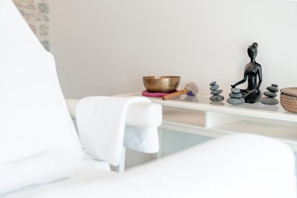 Ein heller Wellnessraum mit gem&uuml;tlicher Liege im Vordergrund. Im Hintergrund befinden sich dekorative Elemente wie eine Buddha-Figur, gestapelte Steine und eine Klangschale, die eine beruhigende Atmosph&auml;re schaffen. Ideal f&uuml;r Erholung und Entspannung.