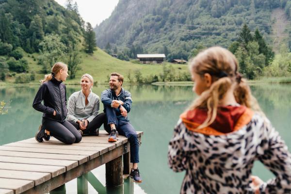 Eine fr&ouml;hliche Familie genie&szlig;t gemeinsame Zeit auf einem Steg an einem klaren Gebirgssee, umgeben von gr&uuml;ner Natur und Bergen. Im Vordergrund steht ein Kind, im Hintergrund entspannen die Eltern und ein weiteres Kind am Ufer. Perfekt f&uuml;r naturnahe Erholung und unvergessliche Momente.