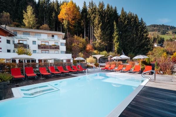 Moderner Außenpool mit hellblauem Wasser, umgeben von roten Liegestühlen und weißen Sonnenschirmen vor einem stilvollen Alpenhotel. Die lebhaften Herbstfarben der umgebenden Bäume sorgen für eine angenehme Wohlfühl-Atmosphäre und laden zum Entspannen ein. Ideal für erholsame Momente inmitten der Natur.